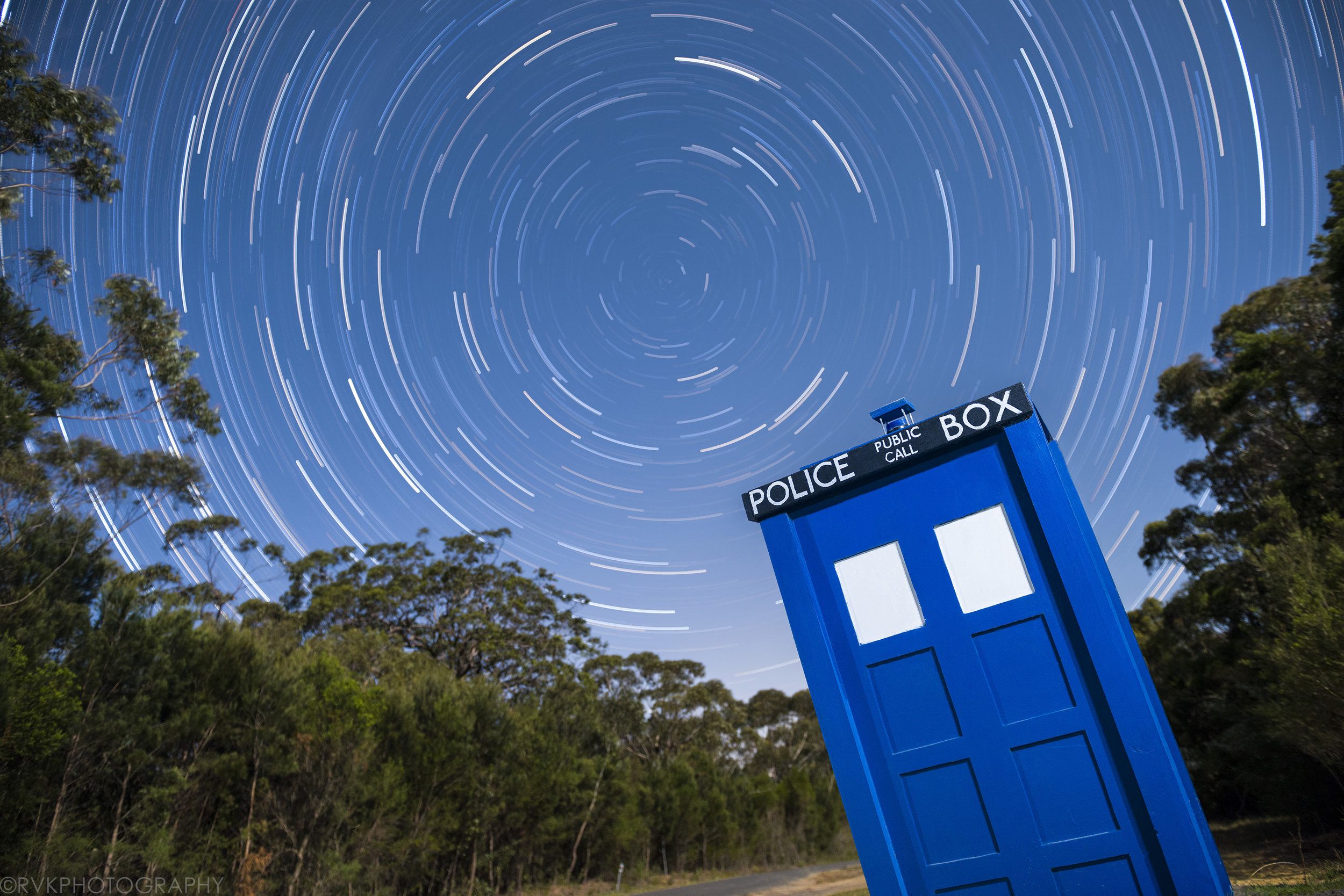The night sky is filled with the swirls of star trails, and the foreground has a bright blue TARDIS police box