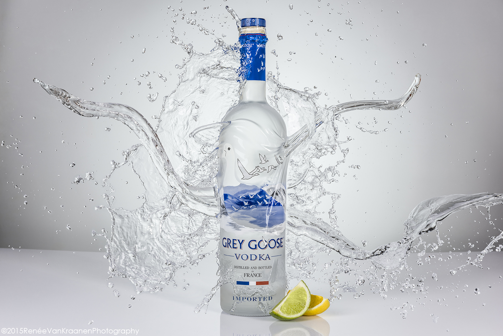 Grey Goose Vodka Product Shot on a white background, there is clear liquid splashing onto the bottle, a wedge of lime and lemon are at the base of the bottle.