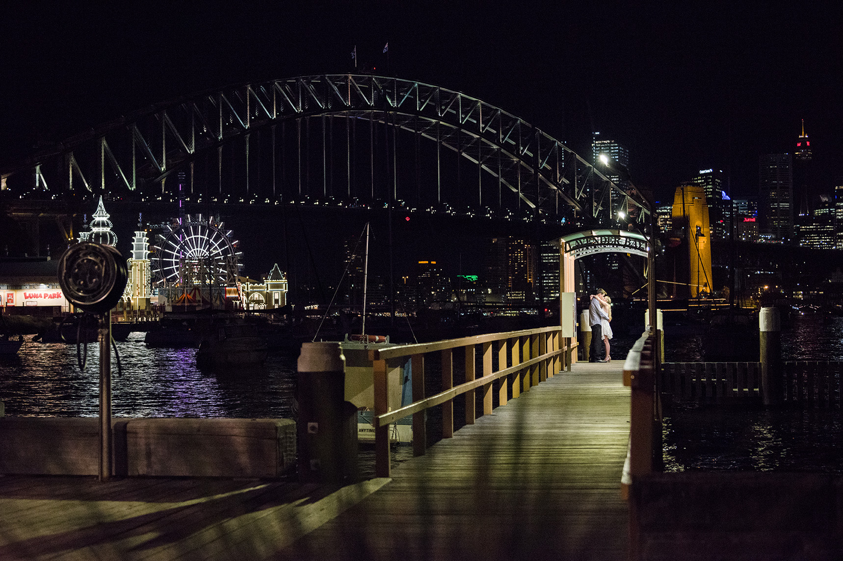 The Sydney Harbour Bridge watches on as two people kiss at the end of a long wharf in the night