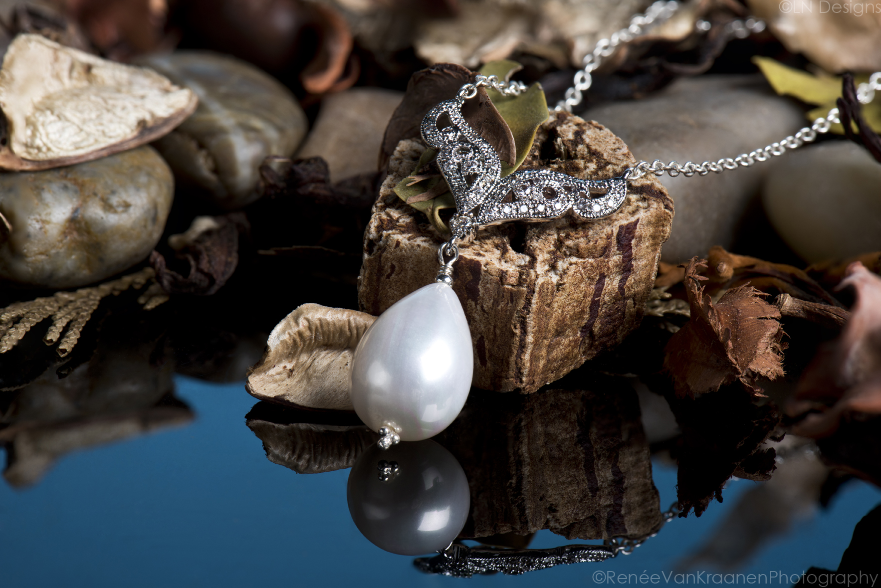 A tear drop pearl necklace sits on some leaves and bark under studio lighting