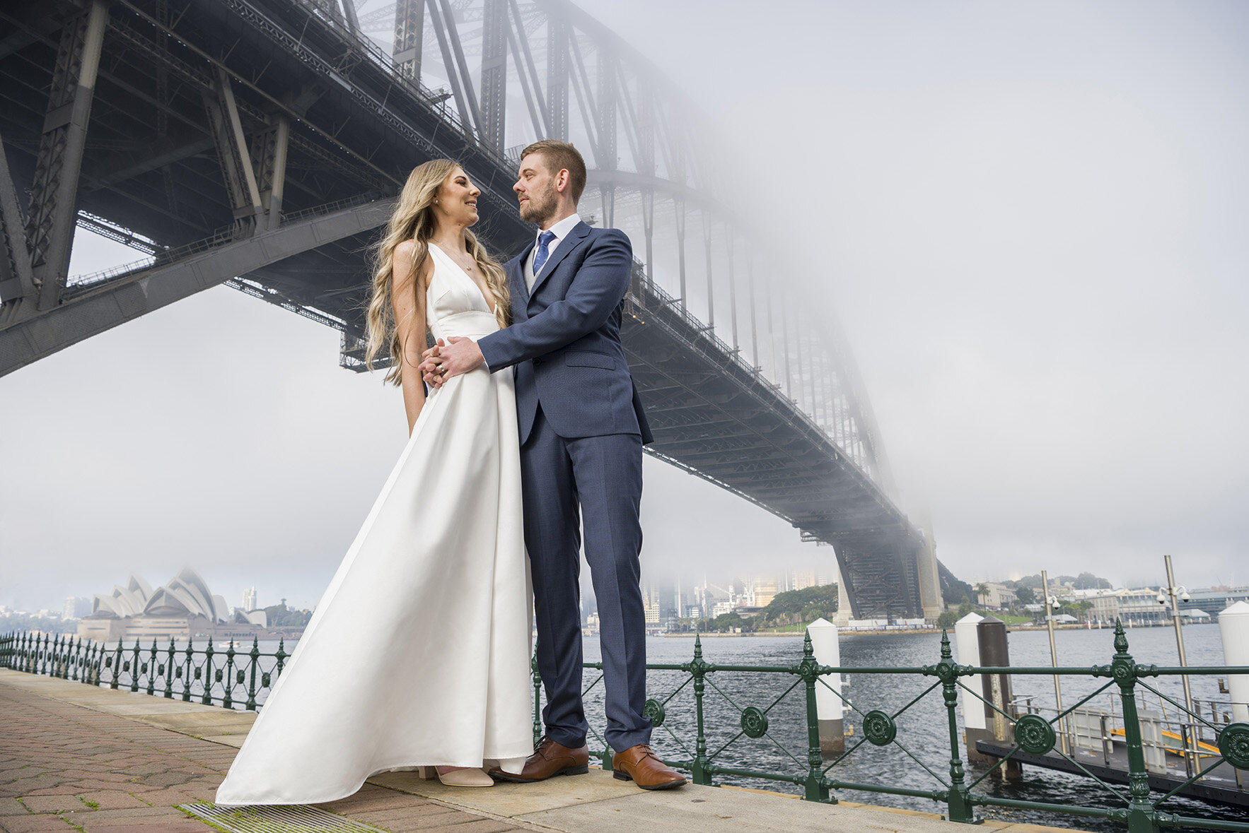 A couple just married wearing a long white dress and blue and silver suite embrace under the Sydney Harbour Bridge on a foggy morning